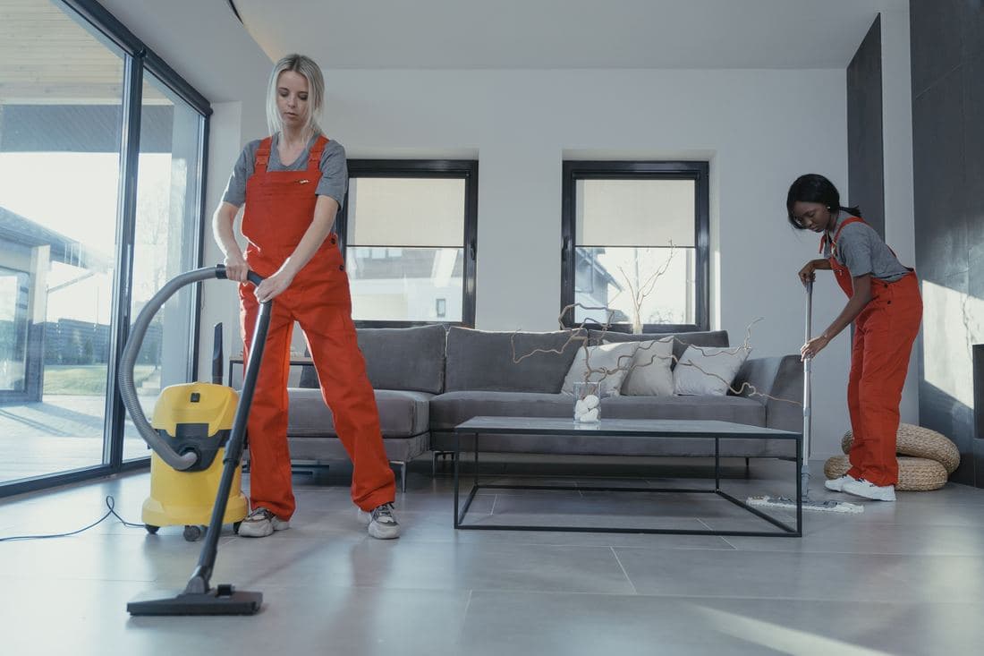 Dos personas de limpieza con uniforme pasando la aspiradora y trapeando una sala